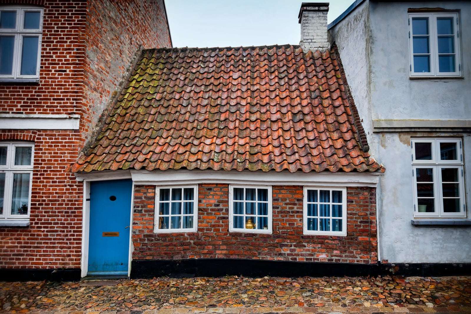 Lille rødt teglhus med tegltag og blå dør mellem to huse