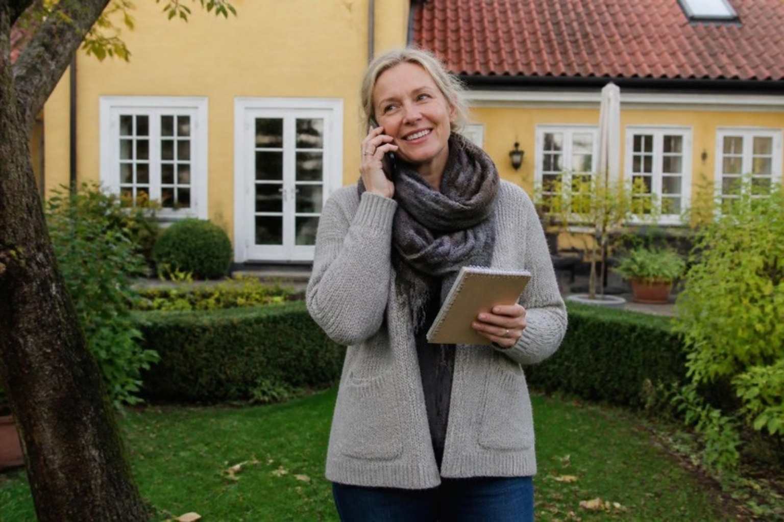 Smilende kvinde med notesbog taler i telefon i haven foran et gulpudset hus med tegltag