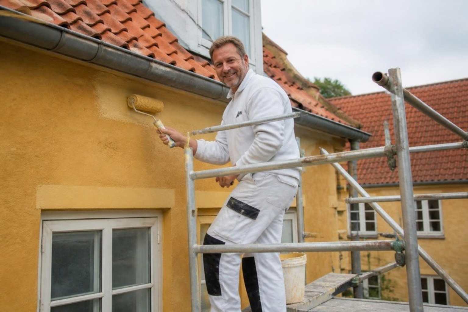 Smilende maler i hvidt arbejdstøj står på et stillads og ruller gul maling på en pudset facade med tegltag